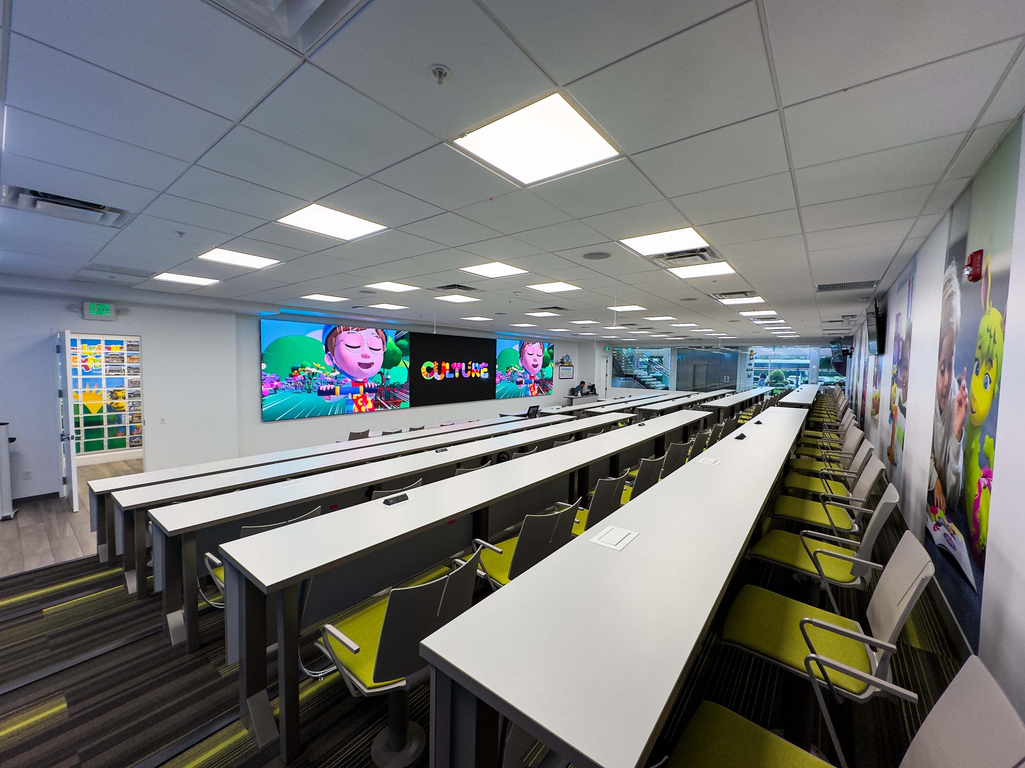 Modern classroom with long white tables, green and gray chairs, and large digital screens featuring colorful animated characters, creating a bright and playful learning environment.