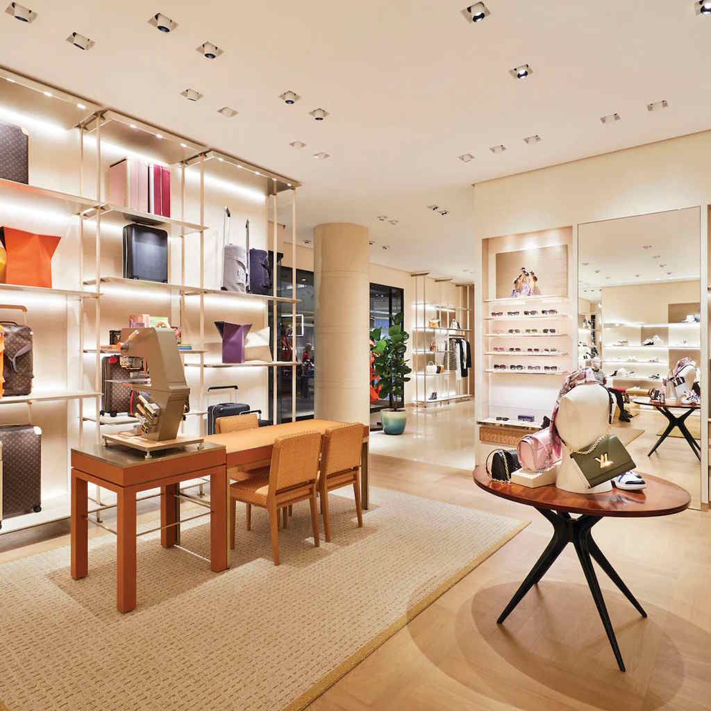 Luxury boutique interior with polished wood floors, minimalist shelving, and curated displays of designer handbags, luggage, and accessories arranged around a central consultation table.