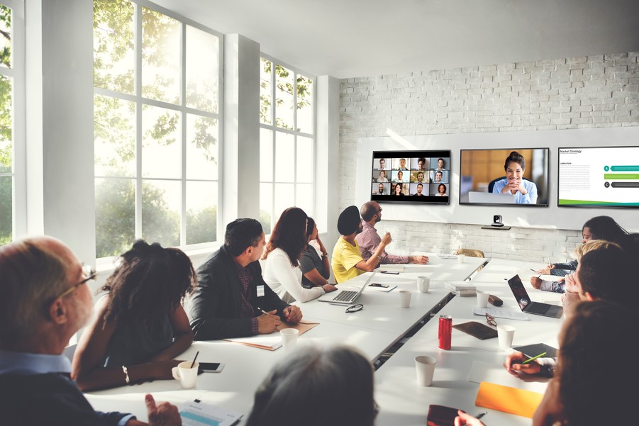 A modern conference room with a long table, people seated around it, and three displays in the front of the room.