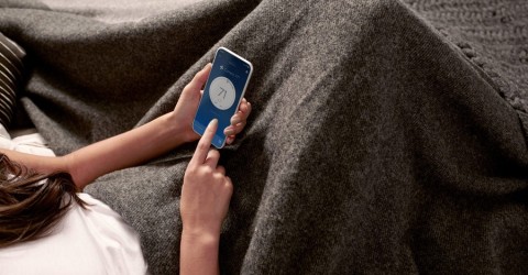 A woman lying under a blanket adjusting the temperature on her phone.