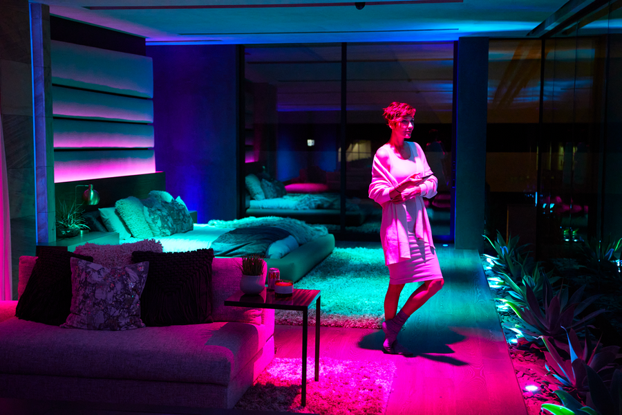 A woman stands in her living room, illuminated by pink, blue, and green hues.