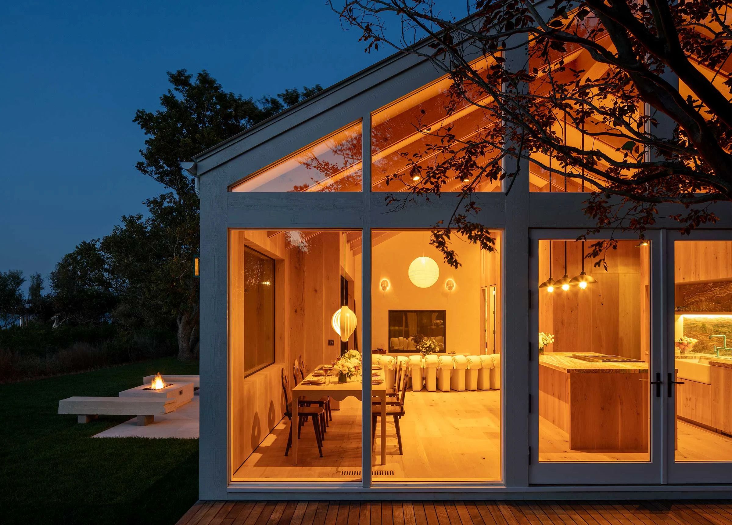 A modern farmhouse style home with picture windows at night with warm lighting inside where a kitchen and dining room are visible though the windows.